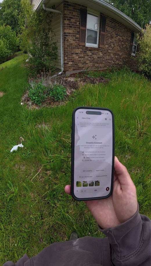 A Belle and Brothers crew member holding an iPhone showing the Grass Go AI Assistant on a real Macomb property